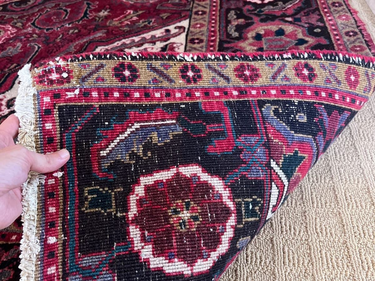 Hand turning back the corner of a hand knotted rug to inspect the foundation, with the fringe and weave visible up close
